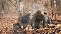 Zwei Soldaten zersägen in einem Wald einen Ast, im Hintergrund sieht man Rauch von einem Lagerfeuer.