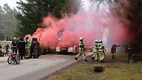 Feuerwehreinsatz auf einer Straße im Wald. Roter Rauch zieht über die Straße. 