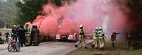 Feuerwehreinsatz auf einer Straße im Wald. Roter Rauch zieht über die Straße. 