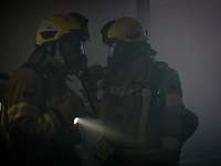 Zwei Feuerwehrmänner mit Atemschutzmasken und einer Taschenlampe in einem verrauchten Raum.