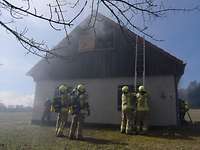 Feuerwehrleute vor brennendem Haus. Zwei Personen stehen im Obergeschoss an einem offenen Fenster.