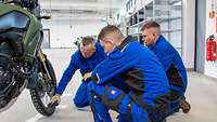 Drei Männer in blauer Arbeitskleidung knien in einer Halle neben einem Motorrad