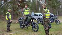 Drei Soldaten in Uniform und mit Warnwesten auf einer Wiese. Einer spricht zu den anderen.