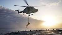 Ein Soldat seilt sich von einem fliegenden Hubschrauber auf das Flugdeck eines Schiffes ab.