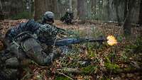 Ein Soldat liegt mit Waffe im Wald auf dem Boden und schießt. Neben ihm liegt ein anderer Soldat.
