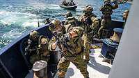 Bewaffnete Soldaten an Deck eines Schiffes. Im Wasser ist ein Schlauchboot mit Soldaten zu sehen.