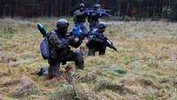 Vier Soldaten stehen und knien mit Waffen auf einer Wiese und sichern sich gegenseitig.