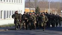 Eine Formation Soldaten marschiert mit Marschgepäck eine asphaltierte Straße hinauf.