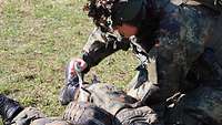 Ein Soldat legt einem Soldaten, der am Boden liegt, einen Verband an.