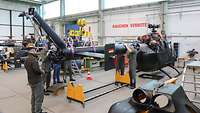In einer Montagehalle stehen mehrere Menschen um einen auseinandergenommenen Hubschrauber.