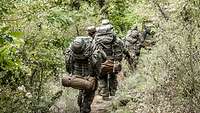 Mehrere Soldaten mit Gepäck marschieren hintereinander einen dicht bewachsenen Waldweg entlang.