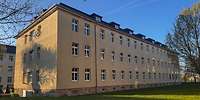 Dreigeschossiger, beigefarbener Häuserblock auf einer Wiese umgeben von Bäumen