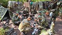 Sechs Soldaten hocken im Kreis und essen. Zwischen Zelten hängt auf Leinen Kleidung.