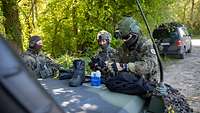 Zwei Soldaten und eine Soldatin stehen an einem militärischen Fahrzeug.