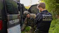 Ein Soldat in Uniform steht mit einem Polizisten vor einem militärischen Fahrzeug und deutet auf eine Skizze