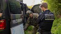 Ein Soldat in Uniform steht mit einem Polizisten vor einem militärischen Fahrzeug und deutet auf eine Skizze