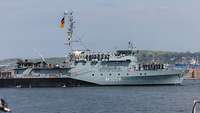 Ein graues Schiff läuft aus mit der Besatzung an Deck.
