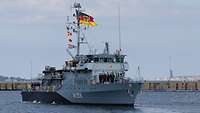 Ein graues Schiff unter einer Deutschlandflagge läuft aus einem Hafen aus.
