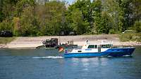 Ein Polizei-Boot auf einem Fluss fährt vor einer militärischen Fähre, auf der sich militärische Fahrzeuge befinden.