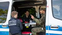 Zwei Polizisten und ein Soldat sitzen in einem Polizeiwagen und sprechen miteinander.