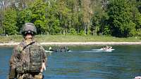 Ein Motorboot fährt auf einem Fluss vor einem militärischen Boot. Im Vordergrund unscharf ein Soldat
