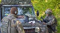 Zwei Soldaten vor einem Geländewagen. Gegenstände wie etwa Schuhe liegen auf der Motorhaube.