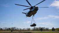 Ein Hubschrauber hebt ein Kettenfahrzeug an einem Haken in die Luft.