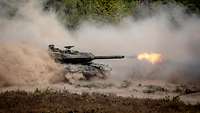 Ein Panzer fährt im Gelände und wirbelt viel Staub auf. Aus dem Kanonenrohr kommt Feuer.