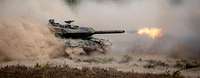 Ein Panzer fährt im Gelände und wirbelt viel Staub auf. Aus dem Kanonenrohr kommt Feuer.
