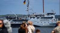 Ein Boot mit Soldaten an Deck fährt in einem Gewässer. Unscharf im Vordergrund sind Personen zu sehen.