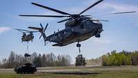 Zwei Hubschrauber fliegen über eine Freifläche. An einem hängt ein kleines Kettenfahrzeug.