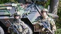 Eine Soldatin und ein Soldat stehen mit ihren Waffen vor einem Militärfahrzeug.