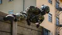 Ein Soldat mit Schutzhelm schwingt sich über eine hohe Holzwand.