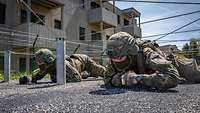 Zwei Soldaten kriechen flach am Boden entlang, knapp über ihnen sind Drähte gespannt.