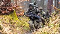 Drei bewaffnete Soldaten laufen hintereinander durch ein Stellungssystem im Wald.