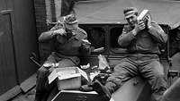 Zwei Soldaten sitzen auf der Motorhaube. Sie essen und hören Musik. Vor ihnen liegt ein Karton.