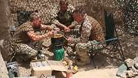 Soldaten sitzen um eine Kochstelle unterm Tarnnetz. Neben ihnen ein Karton und leere Verpackungen.