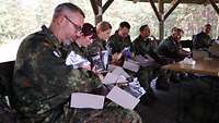 Mehrere Soldaten sitzen in einem Pavillon im Wald und packen Kartons mit Lebensmitteln aus.