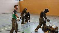 Je zwei Soldaten mit Schutzhelm kämpfen in einer Sporthalle miteinander. Ein Ausbilder leitet an.