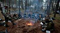 Mehrere Soldaten sitzen im Kreis um eine Feuerstelle im Wald und essen.