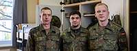 Drei Soldaten stehen nebeneinander vor einem geöffneten Spind und einem Doppelstockbett in einem Raum.