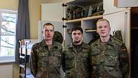 Drei Soldaten stehen nebeneinander vor einem geöffneten Spind und einem Doppelstockbett in einem Raum.