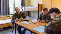 Drei Soldaten schreiben und lesen an einem Tisch in einem Raum. Im Hintergrund stehen Betten und Spind.