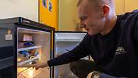 Ein Mann kniet vor einem geöffneten Kühlschrank und nimmt eine Getränkedose heraus.