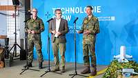 Drei Personen stehen nebeneinander vor einer blauen und sprechen in ein Mikrofon.