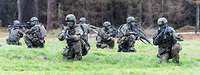 Mehrere Soldaten knien mit ihren Waffen auf einer Wiese vor einem Wald.