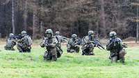 Mehrere Soldaten knien mit ihren Waffen auf einer Wiese vor einem Wald.