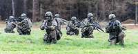 Mehrere Soldaten knien mit ihren Waffen auf einer Wiese vor einem Wald.