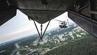 Hinter der geöffneten Heckklappe eines Hubschraubers fliegt ein zweiter Helikopter über einer Stadt.