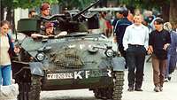 Ein Panzer mit zwei Soldaten fährt durch eine Fußgängerzone, in der viele Menschen spazieren gehen